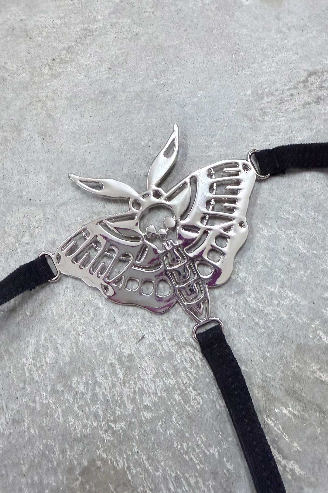 Death Moth Metal Thong - Small featuring a detailed silver moth design with skull and black straps.