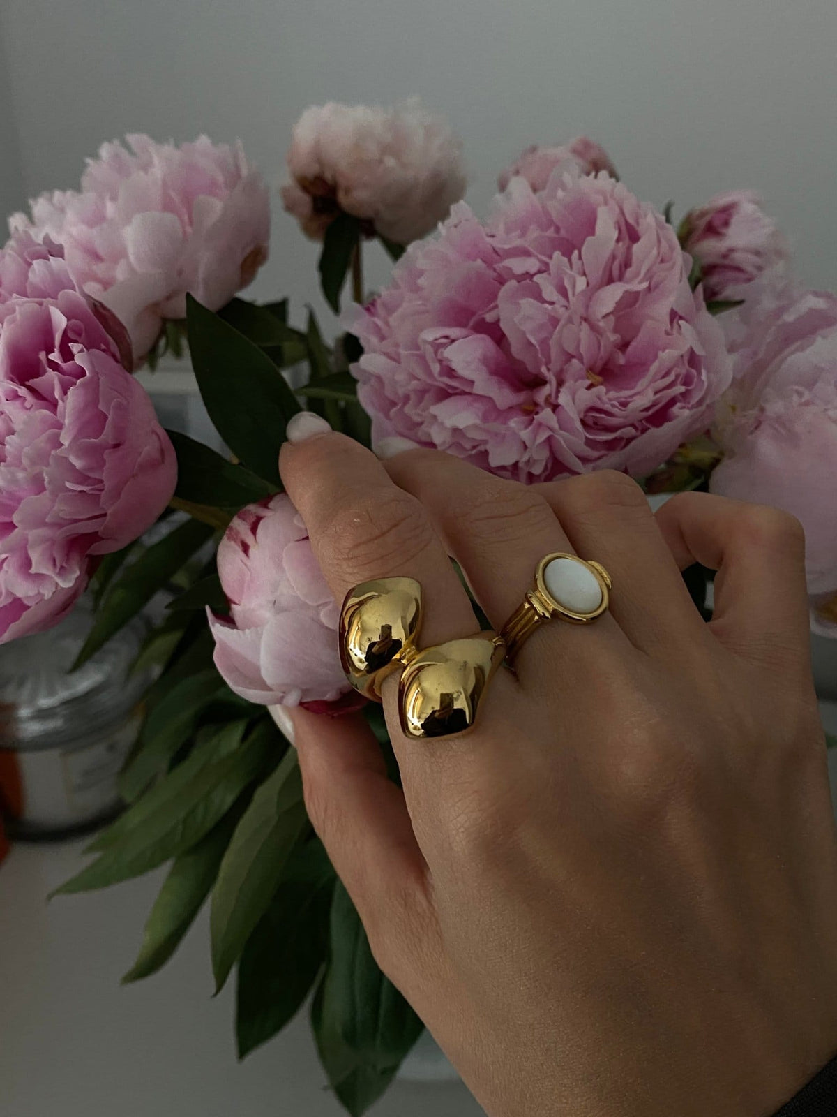 Hand wearing Neva Langais gold ring among blooming pink peonies, showcasing a stylish jewelry collection.