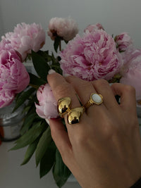 Hand wearing Neva Langais gold ring among blooming pink peonies, showcasing a stylish jewelry collection.